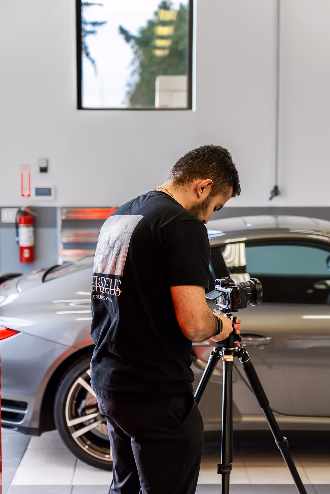 A Perseus videographer on a tripod films a silver sports car inside a dealership, producing cinematic automotive content.