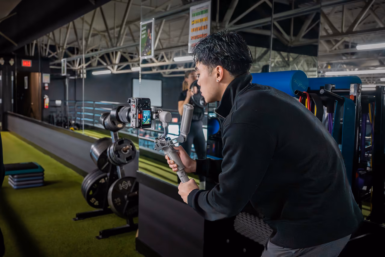 A Perseus Creative Studio videographer crouches with a gimbal-stabilised Sony camera to film fitness content in a gym, framing the weight floor.
