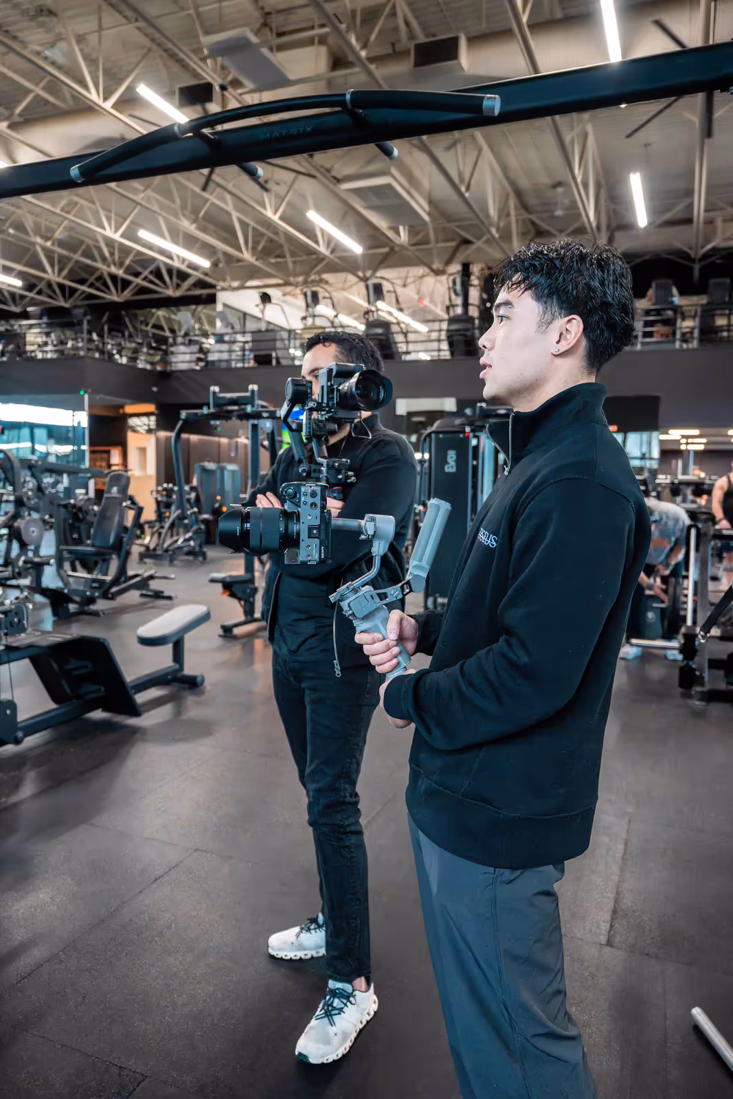 Two Perseus Creative Studio videographers film inside a gym, both operating gimbal-stabilised Sony cameras to capture fitness content.