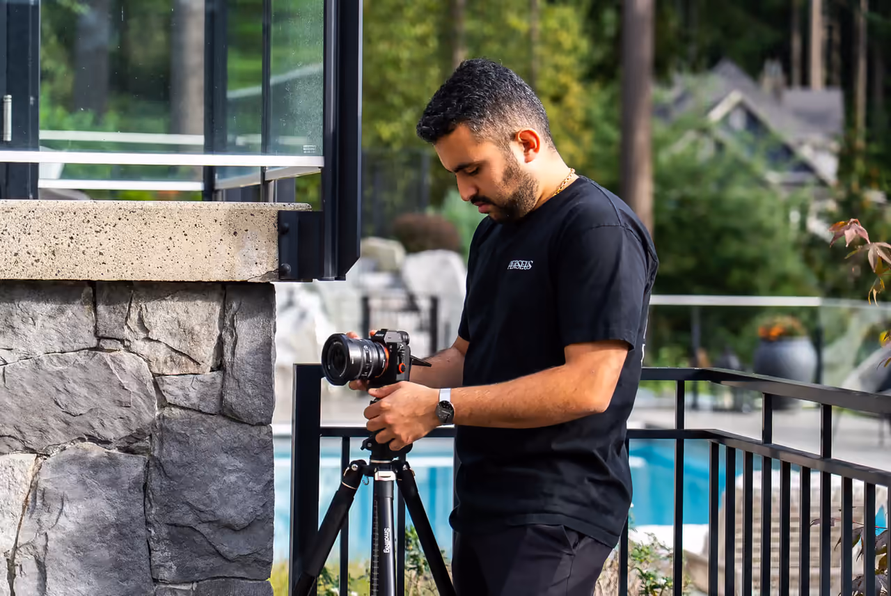 A Perseus Creative Studio videographer films a modern glass-and-stone luxury home and pool from the deck, shooting real-estate content on a tripod outdoors.