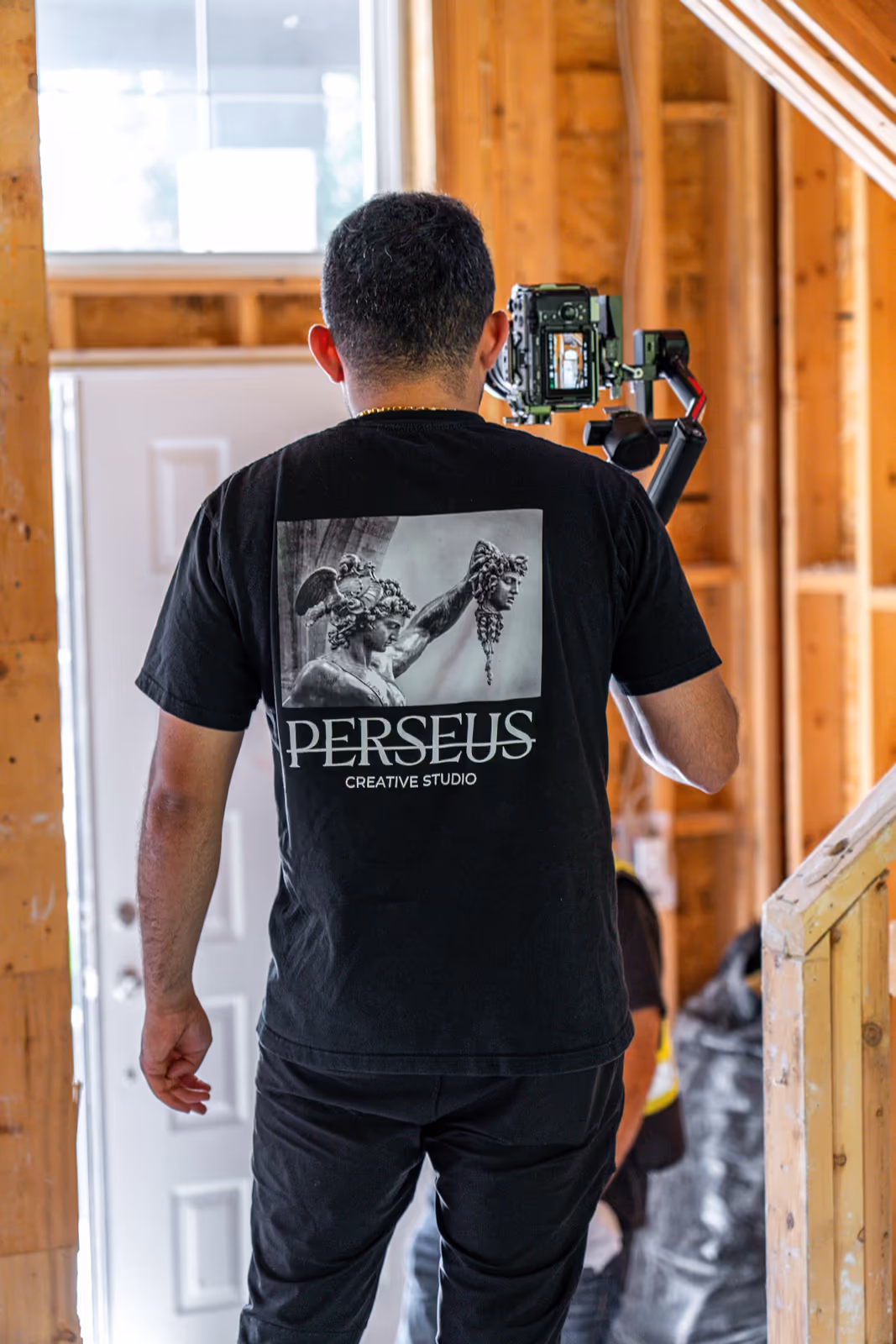 A Perseus Creative Studio videographer in branded apparel carries a gimbal-mounted camera through a doorway while filming on location.
