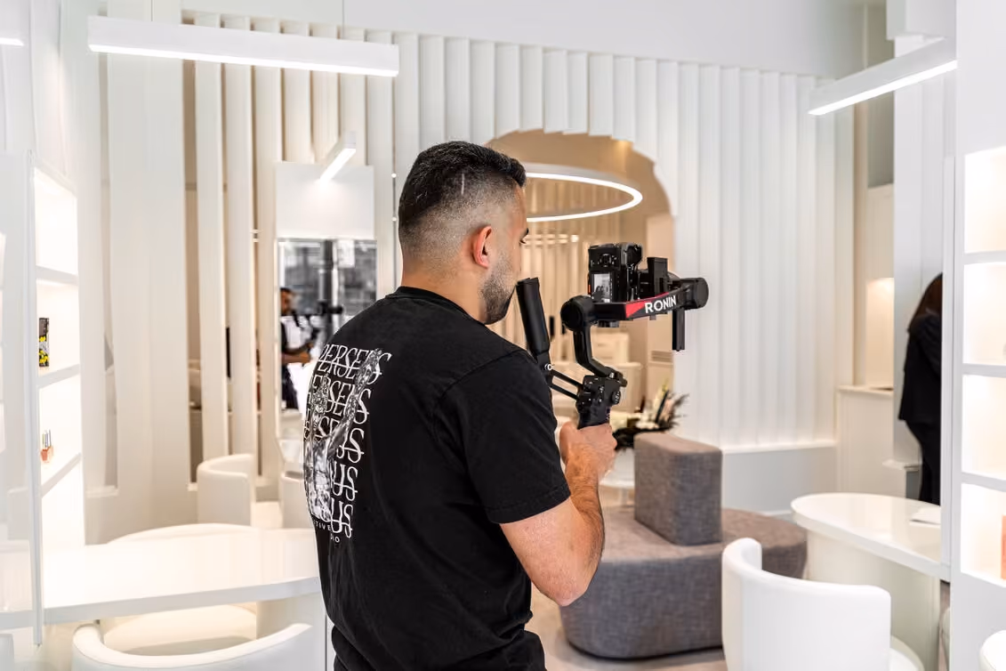 A Perseus Creative Studio videographer, the studio’s Medusa logo across the back of his tee, films with a DJI Ronin-stabilised camera inside a bright, modern boutique interior.