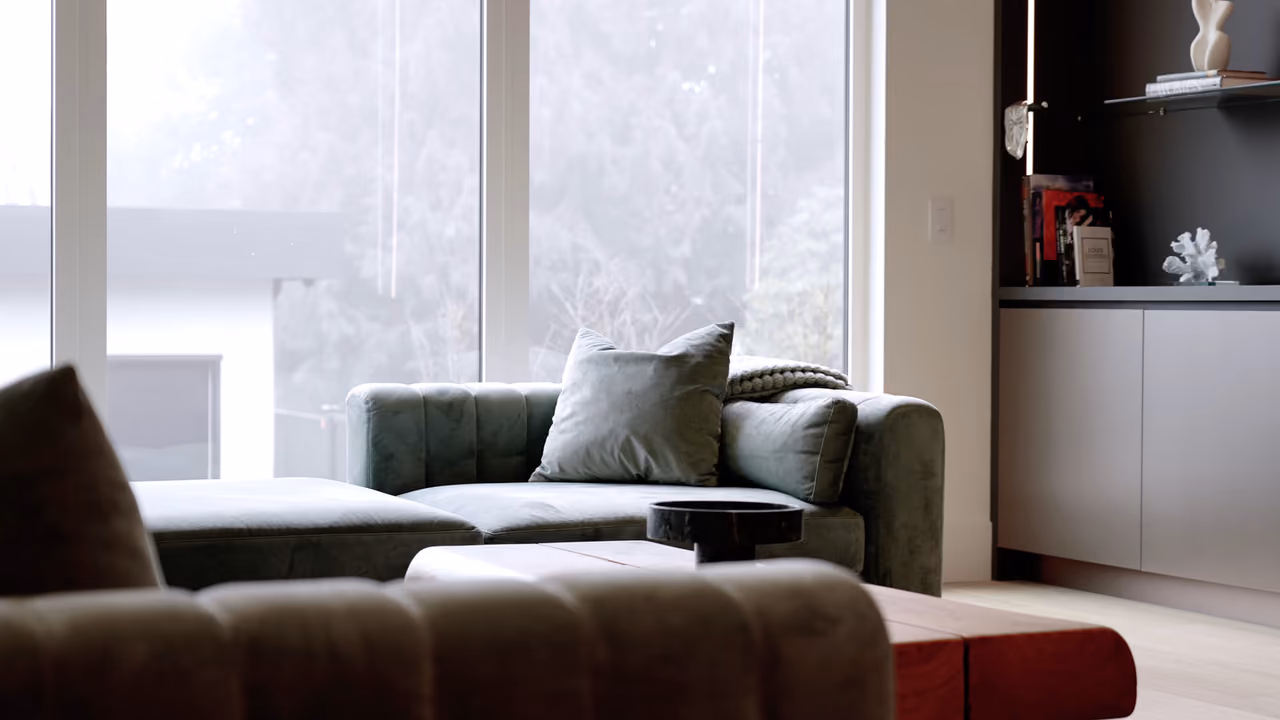 Modern living room with a sage-green velvet sectional and floor-to-ceiling windows in Vela Homes’ West Vancouver custom build on Haywood Ave, filmed by Perseus Creative Studio.