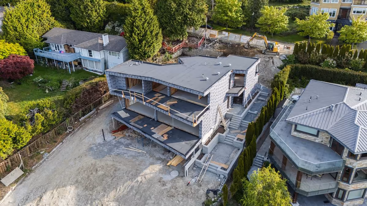 Aerial of a multi-level home mid-construction with sheathing, framed decks, and an excavator on site at Vela Homes’ 1925 Russet Way build, documented by Perseus Creative Studio.