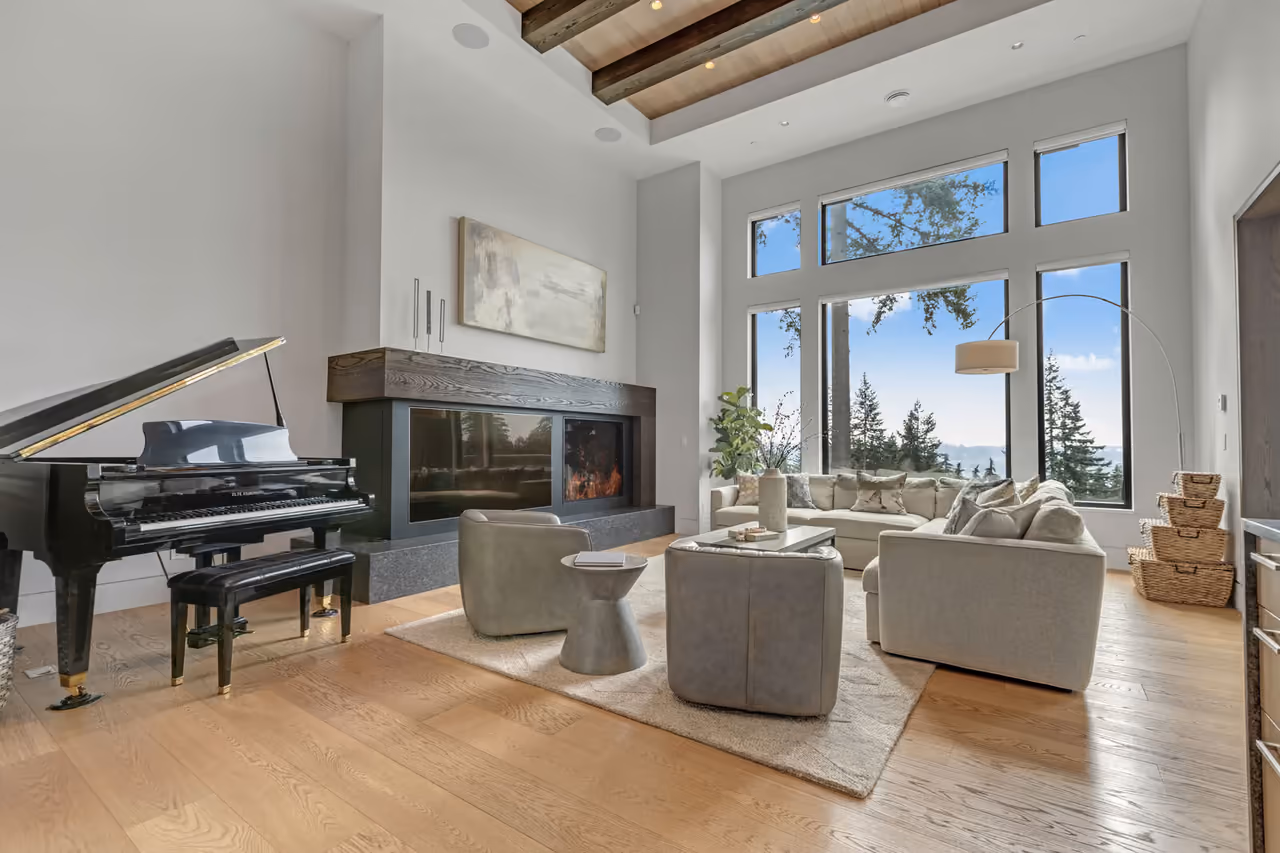 Double-height living room with a grand piano, linear fireplace, and forest-and-water views through floor-to-ceiling windows in a luxury Anmore, BC home, filmed for realtor Erin Price Emery by Perseus Creative Studio.