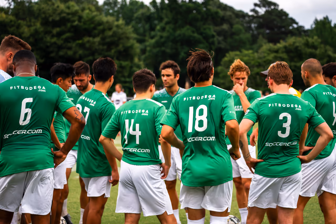 Fitbodega FC players in green kit huddling on an outdoor pitch at the Toronto tournament, captured by Perseus Creative Studio.
