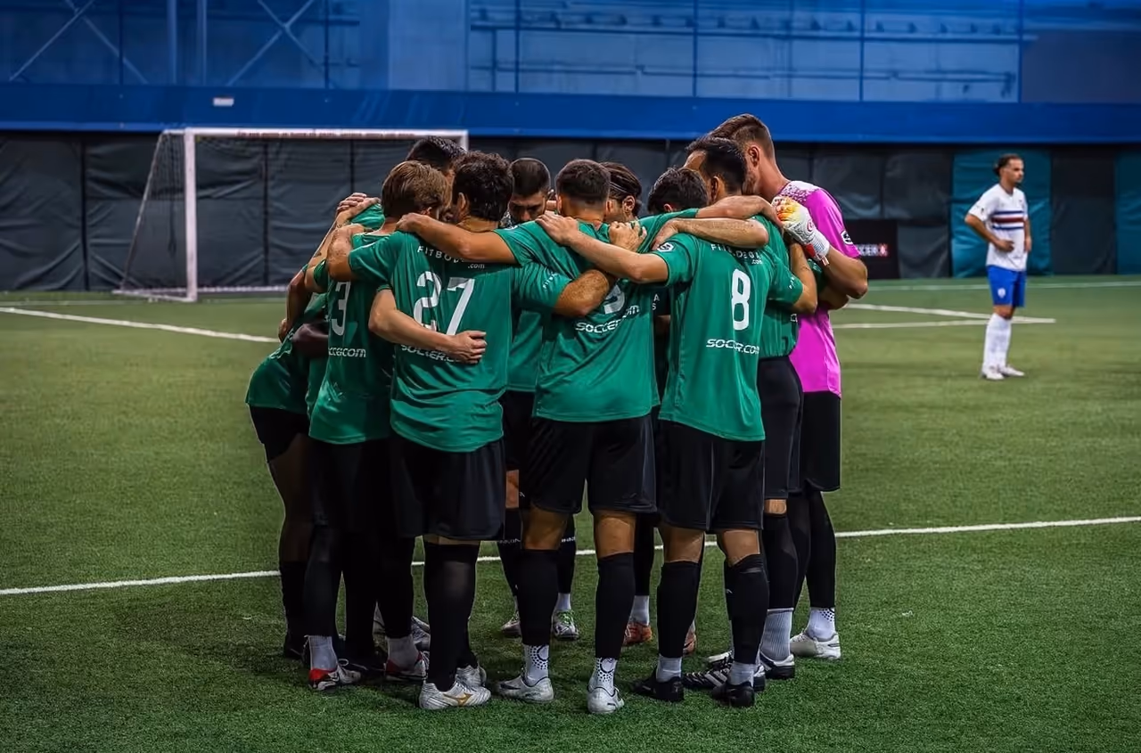 Fitbodega FC team huddle in green kit on an indoor turf pitch during the TST 7v7 trip, photographed by Perseus Creative Studio.