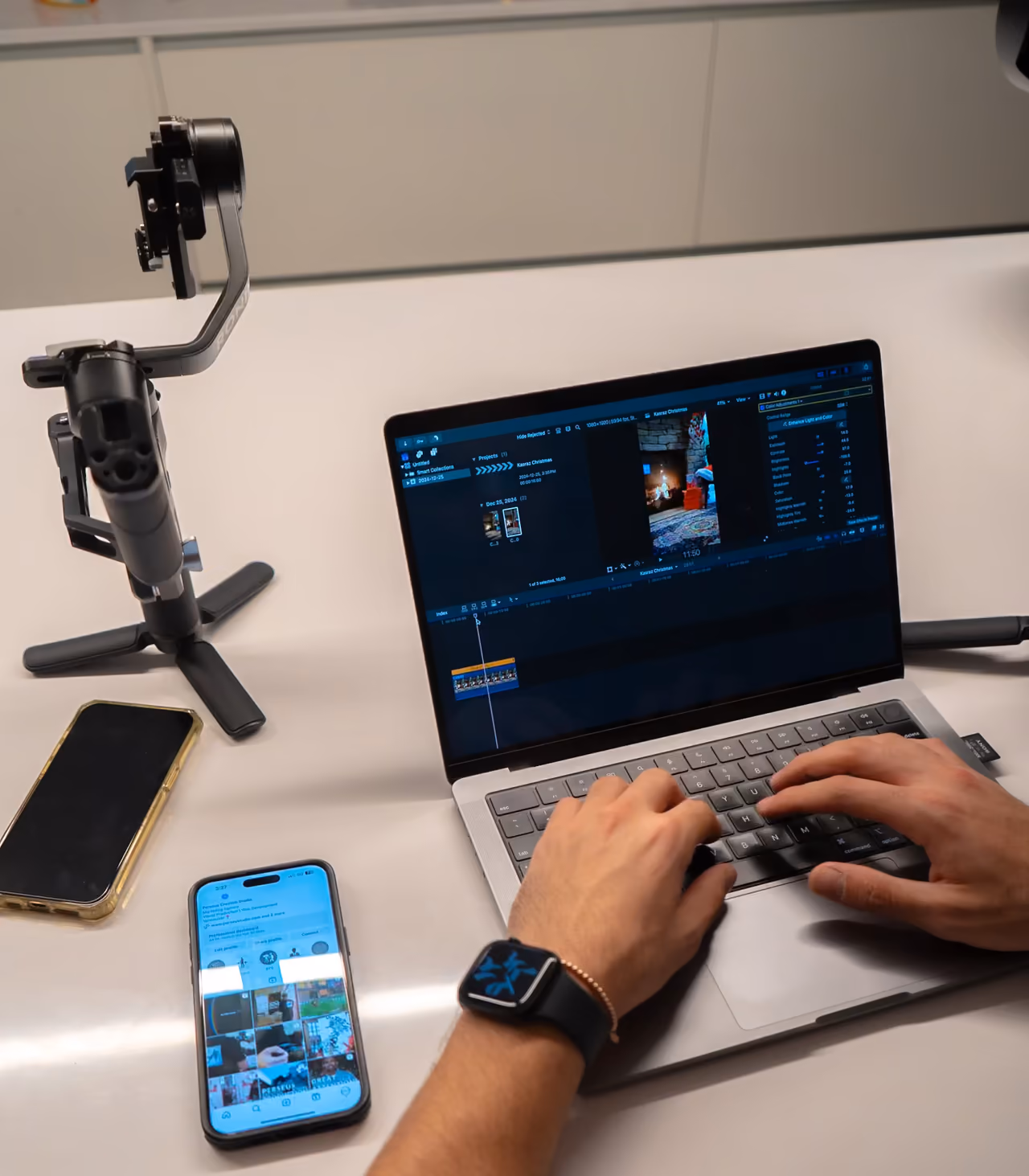 Editor color grading video footage on a laptop editing timeline, with a phone preview and gimbal stabilizer on the desk.